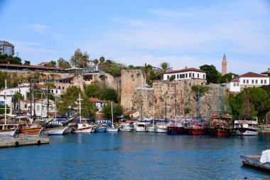 Türkiye 'nin Antalya Limanı' nda demirli tekneler, Kasım 2022. Mavi gökyüzü, su ve arkadaki binalar. Taş şehir duvarları ve minare. 