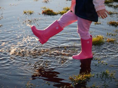 Pembe çizme giyen genç bir çocuk çimenli, güneşli bir alanda sığ bir su birikintisinde su sıçratıyor. Neşeli an, açık havada eğlence, keşif ve kaygısız çocukluk sevincini aktarır..