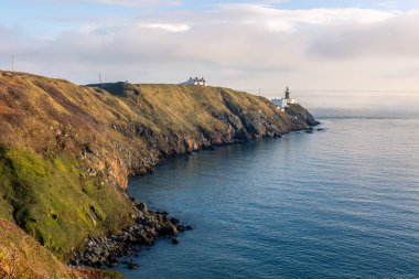 Howth Island, Dublin, İrlanda 'da yürüyüş parkurunda. Uzakta ikonik beyaz Baily Deniz Feneri var. Sakin denizin üzerinde mavi bir gökyüzünün altında sis vardır. Yüksek, dik uçurumlar. Sakin güneşli sisli bir sabah