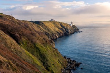 Howth Island, Dublin, İrlanda 'da yürüyüş parkurunda. Uzakta ikonik beyaz Baily Deniz Feneri var. Sakin denizin üzerinde mavi bir gökyüzünün altında sis vardır. Yüksek, dik uçurumlar. Sakin güneşli sisli bir sabah