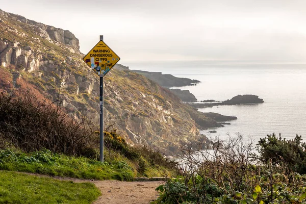Warning sign for steep cliffs on hiking trail on Howth Island, Dublin, Ireland and Baily Lighthouse. Above the calm sea there is fog below a blue sky. High, steep cliffs. Foggy morning in calm sunny