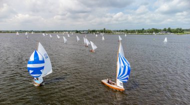 A line of small sailboats in a match on the Sneekermeer, Friesland, The Netherlands. Bright blue and white spinaker sails. White summer clouds and a marina in the background.. High quality photo