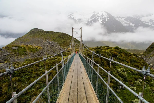 An suspension bridge across an icy stream in Mount Cook National Park, NZ. Hooker Valley track. Cold river, green grass, snow on mountains in background. New Zealand. High quality photo