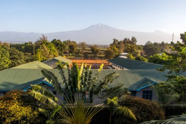 Kilimanjaro Dağı öndeki çalılar ve köyle bulutların arasından görülüyor. Üstü karla kaplı, dramatik bir ruh hali. Yüksek kalite fotoğraf