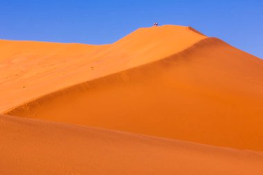 Sossusvlei, Namibya 'da sabah güneşinde gölge dalgalı kırmızı kum tepeleri. Yüksek kırmızı ve turuncu kumulların zarif kıvrımları, mavi gökyüzü. Üstte küçük bir grup yürüyüşçü var. Yüksek kalite fotoğraf