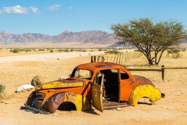Paslı, terk edilmiş araba enkazı. Çölde, Solitaire 'de, Namibya' da, Afrika 'da. Renkli turuncu enkaz, parlak mavi gökyüzünün altında kırmızı pas. Toprak yol, kayalar, akasya ağacı ve kırmızı kumullar. Yüksek kalite fotoğraf