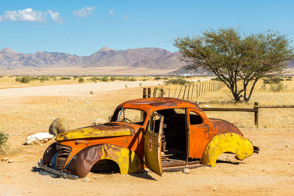Rusty vintage abandoned car wreck in desert, Solitaire, Namibia, Africa. Colourful orange wreck, with red rust under bright blue sky. Dirt road, rocks, acacia tree and red dunes. High quality photo