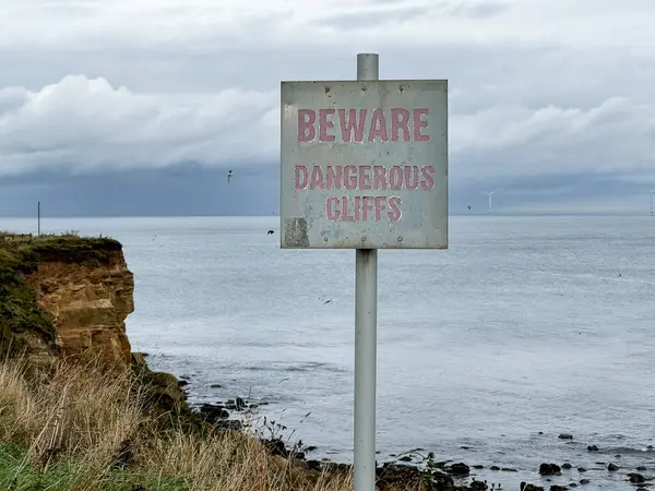 Red sign with white letters saying Beware, dangerous cliffs. In front of eroded cliffs and sea at a beach. In England, UK High quality photo