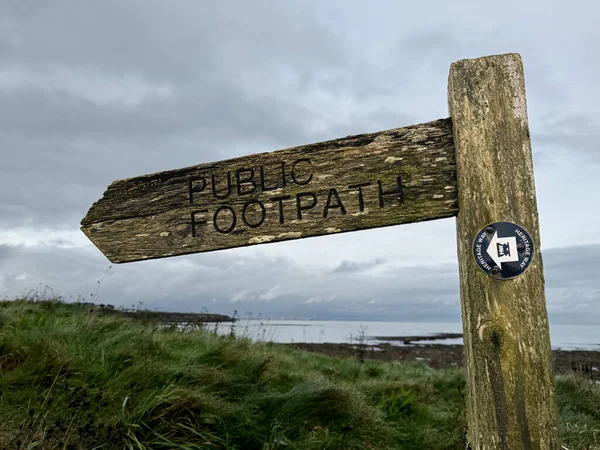 Wooden sign with carved letters saying public footpath. In front of eroded cliffs and the North Sea at a beach. In Whitley Bay, Newcastle, England, UK. High quality photo