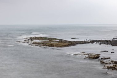 Long exposure of granite stone and rocks along coast coastline in England, uk, Newcastle with blurred waves and grey water of the sea ocean with three fisherman fishing with rod and line.  clipart