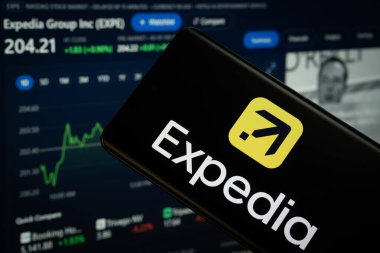 Dhaka, Bangladeş - 08 Mart 2025: Expedia logosu bir akıllı telefon ekranında sergilenmektedir, arka planda bir bilgisayar ekranında bir stok grafik web sayfası görünür.