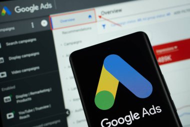 Dhaka, Bangladeş - 16 Mart 2025: Google Ads logosu bir akıllı telefon ekranında sergilenmektedir, arka planda bir bilgisayar ekranında bir stok grafik web sayfası görünür.