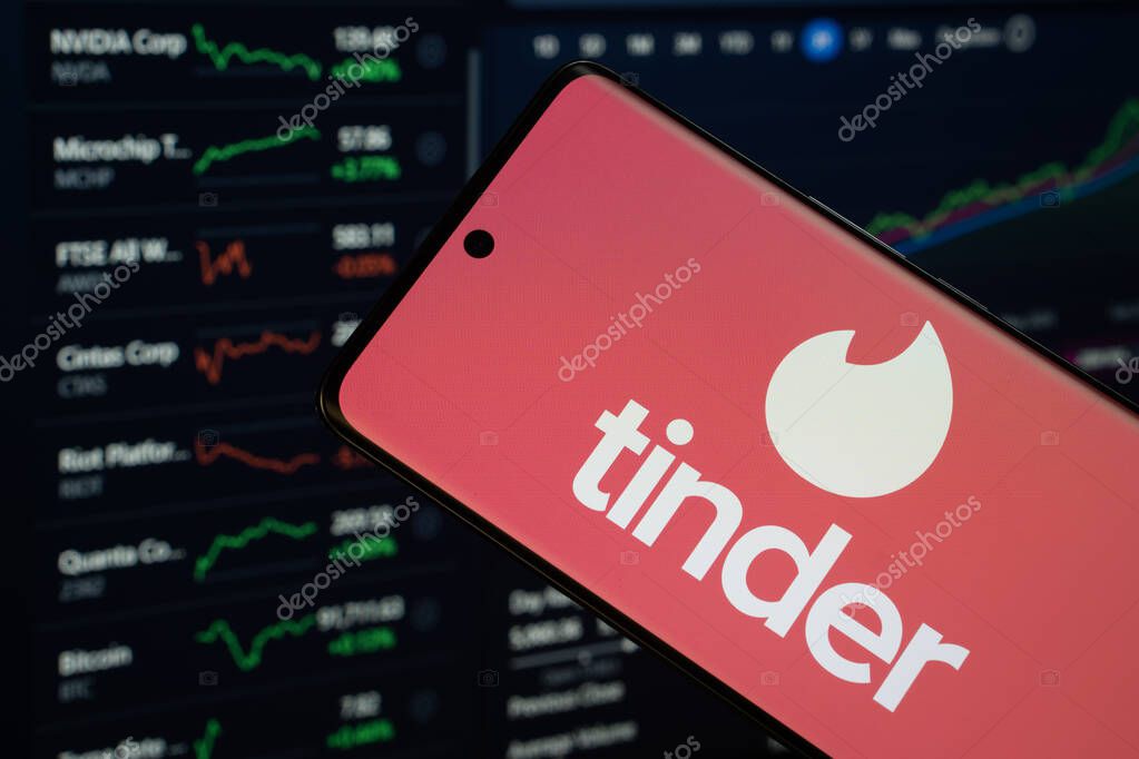 Dhaka, Bangladesh- 18 March 2025: tinder logo is displayed on a smartphone screen, with a stock graph webpage visible on a computer screen in the background.