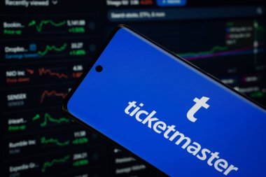 Dhaka, Bangladeş - 18 Mart 2025: Ticketmaster logosu bir akıllı telefon ekranında sergilenmektedir, arka planda bir bilgisayar ekranında bir stok grafik web sayfası görünür.