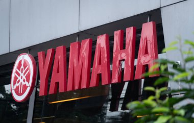 Dhaka, Bangladeş - 17 Ağustos 2025: Kırmızı Yamaha logosu ön planda yeşil yapraklı modern bina cephesinde