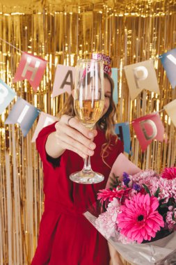 Elinde bir bardak şampanyayla güzel bir kız doğum günü partisinde kadeh kaldırıyor. Kutlama için dekorasyonun arka planında çiçekli sarışın kız.