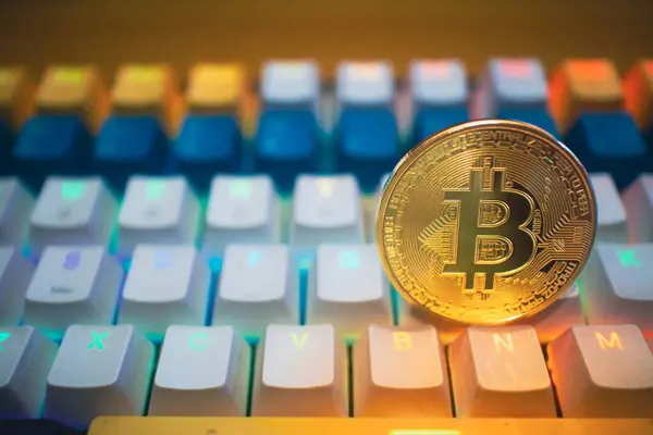 Altın Bitcoin para canlı bir oyun klavyesine yerleştirilmiş renkli ışıklandırma ile dijital para ve modern teknolojinin birleşimini temsil ediyor..