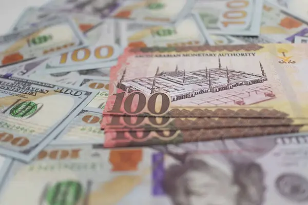Kahverengi Riyal banknotları Amerikan dolarları üzerine istiflenmiş, döviz kurunu, finans piyasalarını ve uluslararası ticari işlemleri sembolize ediyor.