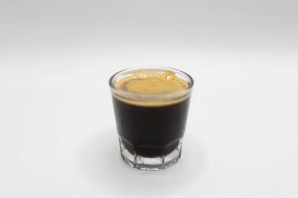 Temiz bir bardakta servis edilen koyu renkli espresso. Pürüzsüz beyaz bir yüzey üzerinde. Kahve severler ve minimalist tasarım görselleri için mükemmel..