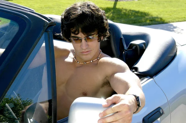 A young hot shirtless and muscular man looks at himself in his rearview side mirror of his sports car to admire his own good looks.
