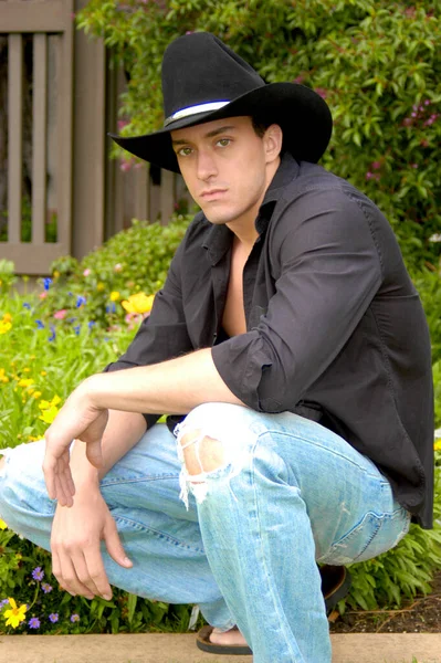 Handsome young cowboy crouches down for a masculine pose with his black cowboy hat.