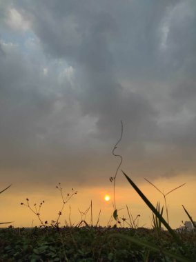 Arka planda güneş batarken bulutlu bir gökyüzü. Gökyüzü birkaç açık mavi parçayla çoğunlukla karanlıktır. Güneş uzaklardan batıyor, manzaranın üzerine sıcak bir parıltı saçıyor.