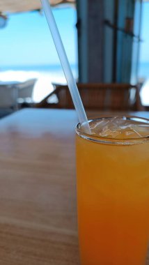 İçinde pipet olan bir bardak portakal suyu. Cam plajın önündeki bir masanın üzerinde.