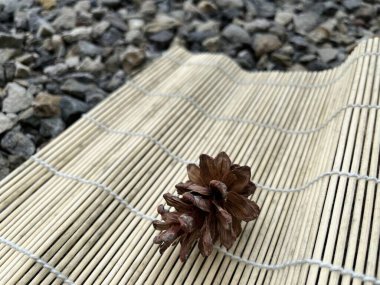 Çam kozalağı, çakıl taşlarıyla çevrili bambu bir hasırın üzerinde dinlenir.