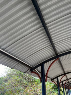 Bu resim siyah kirişler ve kırmızı eğimli desteklerle desteklenen kıvrımlı metal bir çatı içeriyor ve elementlerden korunmak için bir yürüyüş yolu oluşturuyor..