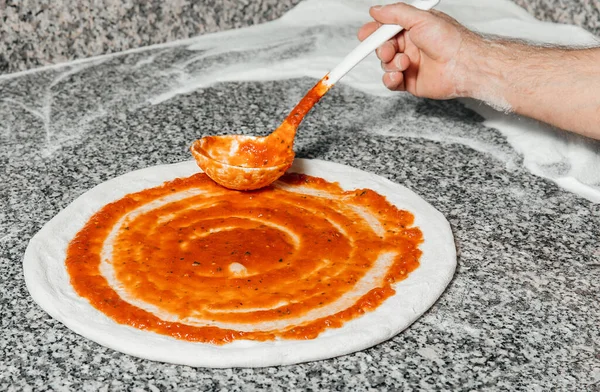 Şef rulo hamurun üzerine domates sosu döküyor, mutfak kaşığı, restoran konsepti, İtalyan pizzası hazırlama süreci.