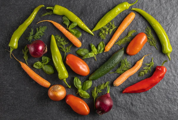 Heart made of fresh seasonal vegetables: green pepper, tomato, onion, carrot, cucumber, basil, dill on grey black background, top view, stock photo