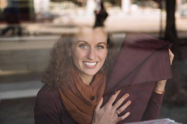 Parlak kıvırcık saçlı bir kadın, bir mağaza penceresinden görülüyor, bir kumaş parçasını tutarken gülümsüyor, neşe dolu bir alışveriş anını gösteriyor.