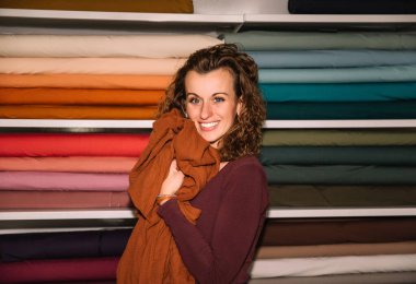  Samimi bir eşarba sarılı gülümseyen bir moda tasarımcısı, kumaş atölyesinde renkli kumaş rulolarının önünde dururken sıcaklığı ve yaratıcılığı yayıyor.