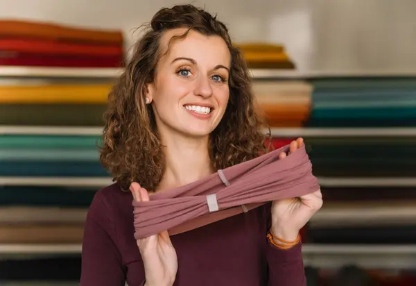 stock image Enthusiastic fashion designer proudly displaying a freshly made textile accessory in her well-organized studio, surrounded by colorful fabric rolls