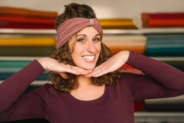 stock image A playful fashion designer showcases her cheerful personality, framing her face with hands while wearing a trendy headband in her vibrant textile studio