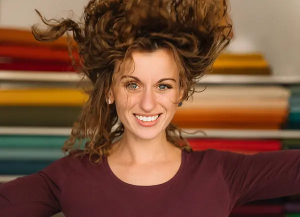 stock image A joyful young woman with lively curly hair playfully poses in her colorful fabric shop, radiating happiness and vitality