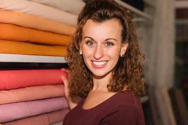 stock image Young woman with curly hair and a bright smile choosing fabric rolls in a vibrant craft store