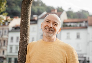 Mutlu kıdemli adam sarı bir kazak giyiyor, mutlu görünüyor ve gülümsüyor, güneşli bir şehir ortamında tarihi binalarla birlikte.