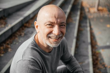 Yaşlı adam merdivenlerde oturuyor, kameraya gülümsüyor, sonbahar yaprakları etrafa saçılıyor.