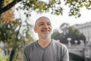 Yaşlı adam parkta, ağaçlarda ve nehirde otururken sıcak bir şekilde gülümsüyor.