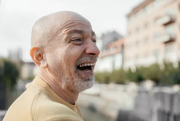 stock image Elderly man in a yellow sweater laughing joyfully, capturing a moment of happiness outdoors with an urban landscape backdrop