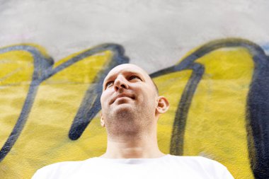 Beyaz tişörtlü kel adam yukarı bakıyor, duvar arkası grafiti, sarı ve siyah renkler.