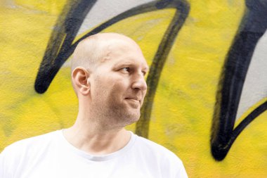 Beyaz tişörtlü kel adam yan bakıyor, renkli grafiti arka planı, şehir merkezi.