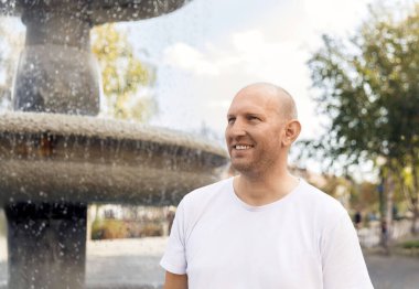 Beyaz tişörtlü kel adam çeşmenin yanında gülümsüyor, açık hava parkı, güneşli bir gün.