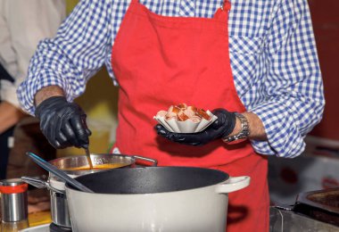 Chef dressed in a red apron cooking, holding sausage slices, plated ingredients, preparing a delicious dish in a professional kitchen, culinary arts concept clipart