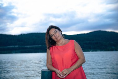 Kırmızı elbiseli bir kadın bulutlu bir gökyüzü ile gölün kenarında duruyor, sakin bir ifade sergiliyor; su manzaralı doğa arka planı