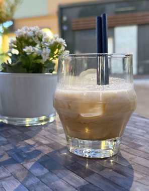 Açık hava çiçeğinin yanındaki desenli masada siyah pipetli buzlu kahve.