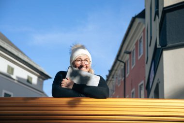 Smiling woman in winter attire with laptop outdoors on a sunny d clipart