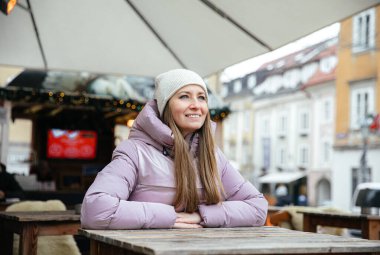 Kışlık ceketli gülümseyen kadın şehir merkezinde rahat bir kafede oturuyor.