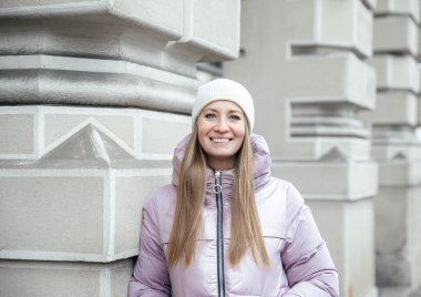 Kışlık ceketli ve bereli gülümseyen kadın mimari duvara yaslanıyor.